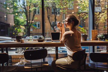 Caucasian romantic woman blogger relaxing drinking tea while sitting near the window in modern loft cafe bar,female freelancer thinking about new ideas during work on laptop computer. Copy space