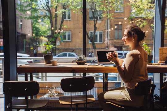 Caucasian Romantic Woman Blogger Relaxing Drinking Tea While Sitting Near The Window In Modern Loft Cafe Bar,female Freelancer Thinking About New Ideas During Work On Laptop Computer. Copy Space