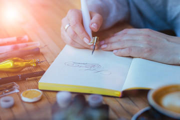 Close up of hands woman calligrapher writes something on notepad. Inscribing ornamental decorated letters. Calligraphy, lettering, graphic design handwriting, creation concept. Sunglare effect.