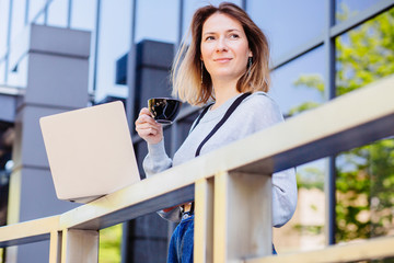 Business female student dreamer freelancer standing outside of cafe holding coffee cup and laptop and looking aside over modern urban background. Close up portrait