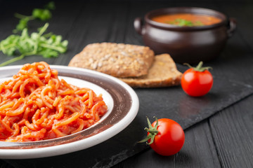 Vermicelli in tomato sauce. Nearby are cherry tomatoes, two slices of bread, and there is a wax soup in the background.
