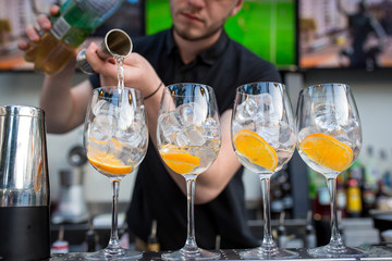 four glasses standing on the bar. Glasses are with ice and orange. Barman pours alcohol in glasses from a measuring glass
