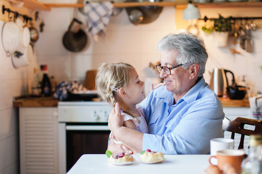 Happy grandmother is hugging granddaughter in cozy home kitchen. Senior woman and cute little child girl are smiling. Kid is enjoying kindness, warm hands, care, support. Family is cooking together.