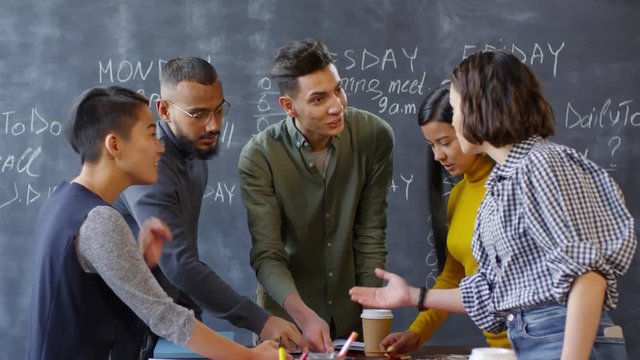 Thigh-up shot of five 20-something multiethnic creative professionals standing at table in front of chalkboard and enthusiastically discussing project workflow chart, talking all at once and smiling