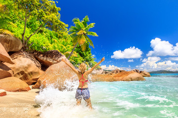 Funny tourist woman in splashing wave at Felicite Island, La Digue. Tropical jungle landscape of Seychelles. Granite boulder stone and waves crashing on shore. Leisure female in bikini at tropics.
