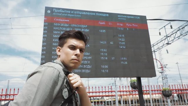 Mineralnye Vody, Stavropol Territory, country Russia 05/24/2019. A young man stands on the platform of the railway station at the board with the train schedule and looks at the screen of the phone, wh