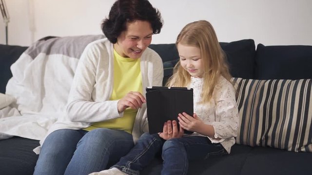 Laughing senior grandmother and cute grandchild is watching funny video on tablet. Family is sitting on sofa and talking, using digital technology tablet device. Web generation and social game showing