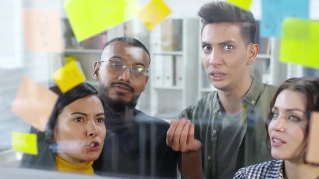 Close-up shot of four young multiethnic colleagues standing in front of glass wall in creative studio and looking at colourful memos, animatedly discussing project tasks and objectives