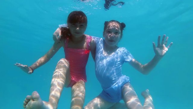 Two cute little girls hug and pose for the camera underwater at the bottom of the pool in a pink and blue swimsuit on a sunny day. They look at the camera, smile and wave their hands. Slow motion. 4K