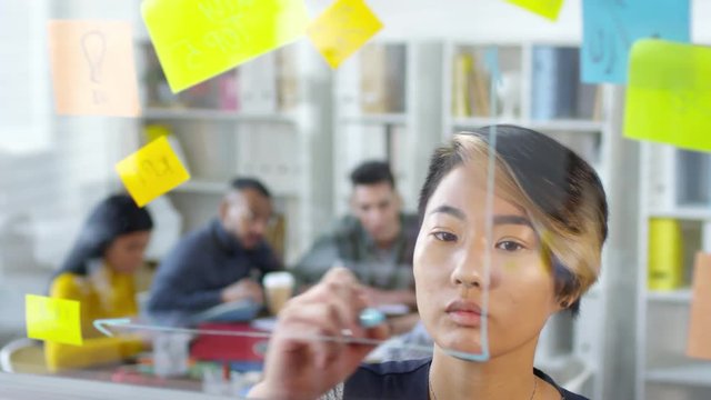 Close-up shot of focused 20-something Asian woman standing in front of glass partition with colourful memos and drawing graph with blue marker, while her colleagues are having meeting in background