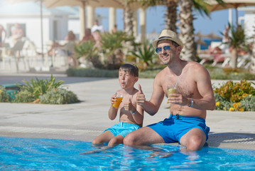Fototapeta na wymiar Dad and son are sitting on the side of the swimming pool, drinking lemonade and making like gestures. They are at the summer holidays.