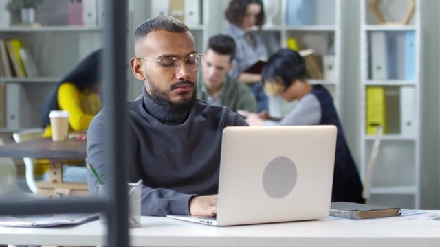 Panning waist-up shot of focused African American colleague sitting at desk and working on laptop in informal office, with other team members busy in background and someone else walking by