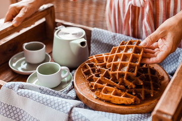 Women holds fresh baked waffles and tea mugs for breakfast. Morning coffee with homemade cakes. Toned photo. Cozy atmosphere at home and flowers on the table.