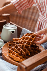 Women holds fresh baked waffles and tea mugs for breakfast. Morning coffee with homemade cakes. Toned photo. Cozy atmosphere at home and flowers on the table.