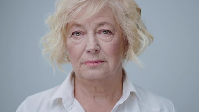 Portrait of nice aged blonde model with green eyes isolated on grey background. Beautiful mature woman with serious expression in white shirt looking at camera.