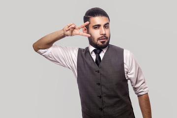 Portrait of funny handsome bearded brunette man in white shirt and waistcoat standing with victory, peace sign and looking at camera. indoor studio shot isolated on gray background.