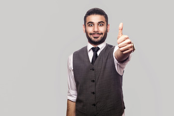 Portrait of happy satisfied handsome bearded brunette man in white shirt and waistcoat standing, smiling and looking at camera with thumbs up. indoor studio shot isolated on gray background.