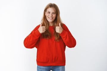 Obraz na płótnie Canvas Portrait of charming young girl tottally agrees smiling broadly showing thumbs up gesture to give positive reply, liking good idea, cheering, being supportive standing in warm cozy red sweater