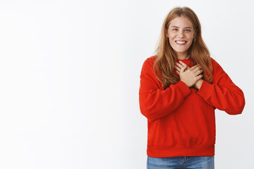 Pleased happy touched young girl with red hair, freckles and blue eyes smiling joyfully with white teeth pressing palms to heart melting from positive compliments, being thankful over grey wall