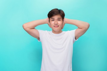 Too loud. Frustrated young man covering ears with hands and looking at camera while standing blue background