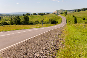 Fototapeta na wymiar Asphalt road going across mountains and green forests. Trees and their shadows on the grass. Sunny summer day with blue sky. Ural landscape
