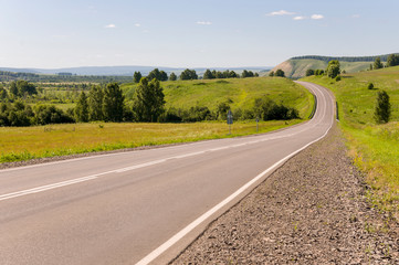 Fototapeta premium Asphalt road going across mountains and green forests. Trees and their shadows on the grass. Sunny summer day with blue sky. Ural landscape