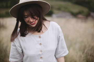 Stylish girl in linen dress and hat walking among herbs and wildflowers in field. Boho woman smiling and relaxing in countryside, simple slow life style. Space for text. Atmospheric image