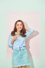 Glad delightful young asian woman with toothy smile looks at camera, holds tasty ice cream, stands on light pink background in tourist outfit, glasses