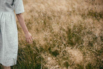 Stylish girl in linen dress holding hand among herbs and wildflowers in field. Boho woman walking in countryside among grass, simple slow life style. Space for text. Atmospheric image