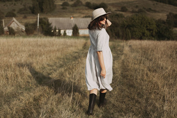 Stylish girl in linen dress and hat running and smiling in sunny field grass at village  in mountains. Boho woman relaxing in countryside, simple rustic life. Atmospheric image. Space text