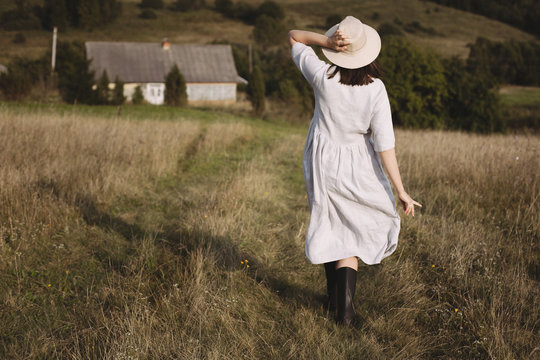 Stylish girl in linen dress and hat walking in sunny field grass at village  in mountains. Boho woman relaxing in countryside, simple rustic life. Atmospheric image. Space text