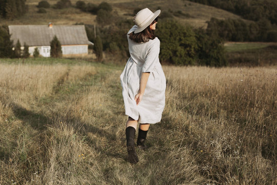 Stylish girl in linen dress and hat running and smiling in sunny field grass at village  in mountains. Boho woman relaxing in countryside, simple rustic life. Atmospheric image. Space text