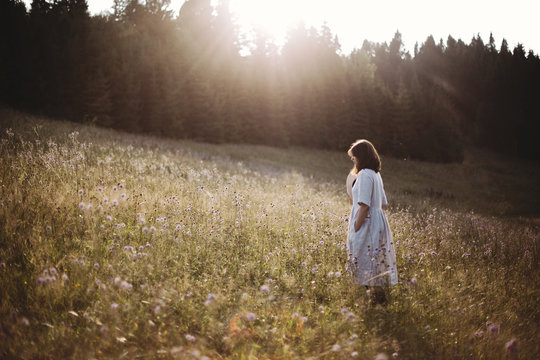Stylish girl in linen dress walking among wildflowers in sunny meadow in mountains. Boho woman relaxing in countryside flowers at sunset, simple life. Atmospheric image. Space text