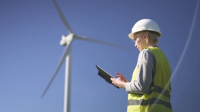 Adult or mature collar worker in white hard hat and green uniform wear standing with modern tablet against wind farm power station on beautiful landscape. Female using program on device