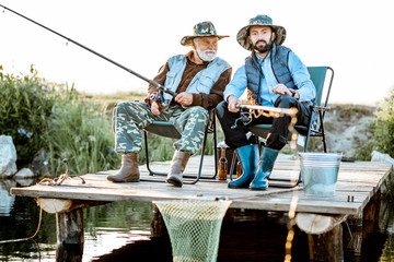 Grandfather with adult son fishing together on the wooden pier during the morning light. View from the side of the lake