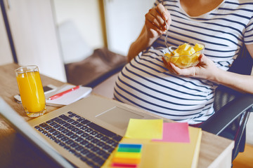 Young Caucasian pregnant freelancer eating fruits and sitting at home office. On desk laptop, glass with fresh orange juice and notebooks. Working pregnant women concept.