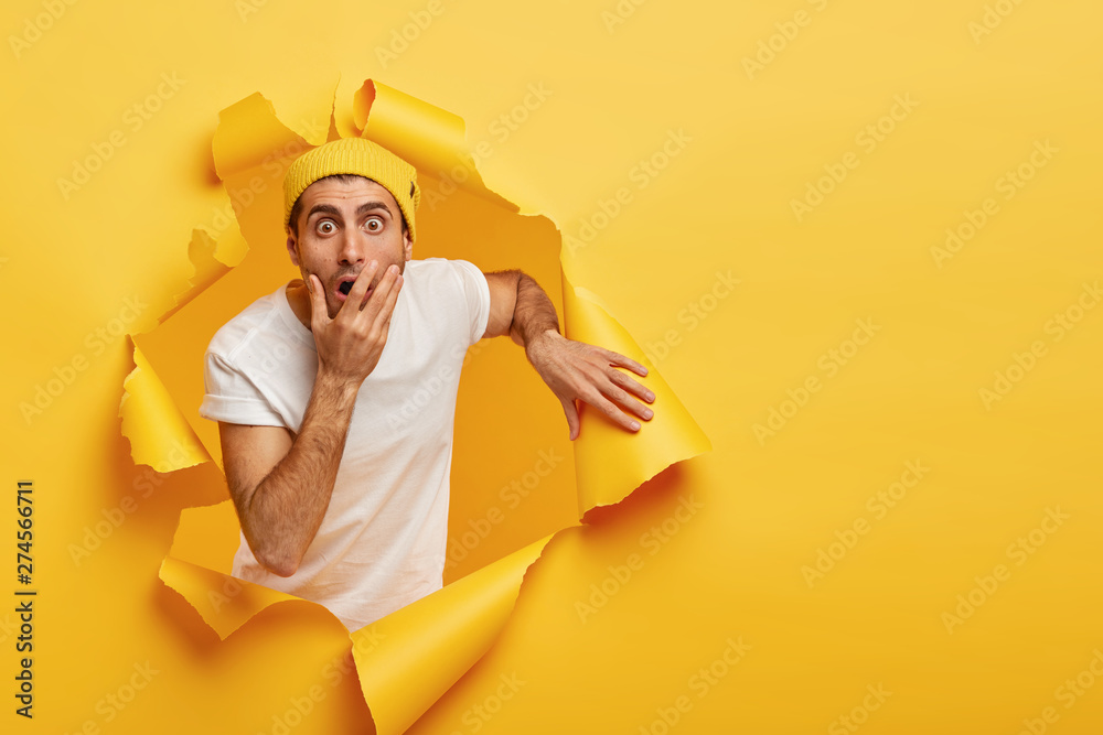 Wall mural image of frightened puzzled guy covers mouth, stares with scared expression, cannot believe his eyes - Wall murals