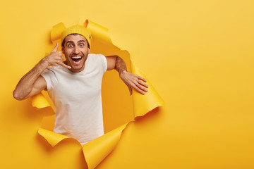 Young positive attractive male model makes call gesture, wears yellow hat and casual white t shirt,...