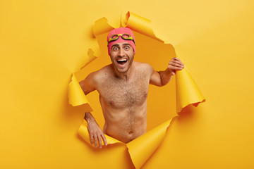 Overemotive man swimmer poses shirtless, wears swimhat and goggles, poses in torn paper hole,...