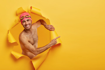 Handsome cheerful man recreats during summer hoidays, makes creative photo, poses in ripped torn paper hole, uses swim equipment, indicates on free space, shows place for good rest on vacation