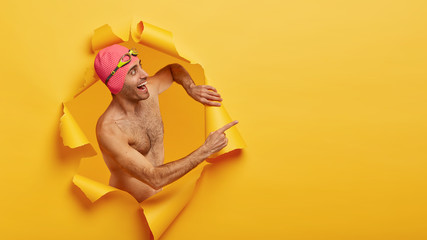 Happy professional male swimmer recreats in swimming pool, wears pink swimcap with goggles on head,...