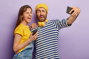 Positive guy and female blink eyes, pose in front of cell phone camera, make photo for internet blog, take selfie, have glad expressions, wear casual outfit, enjoy leisure time. Bloggers with gadgets