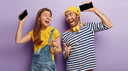 Millennial happy girl and man hold cellphones, dance to music, have fun together, look joyfully at each other, enjoy fantastic sound, listen music using headphones, isolated on purple background
