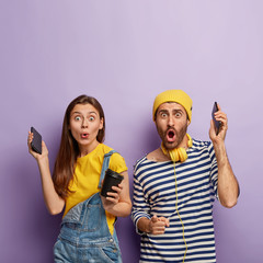 Happy surprised nice couple have fun together, raise hands with smartphones, hear pleasant music, drink coffee, enjoy free time, dance against purple wall, wear striped jumper, hat, denim overalls