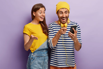 Optimistic young female and male users of smart technology, feel good from successful cellphone updating, gaze at screen, check article, wear casual clothes, stand closely over purple background.