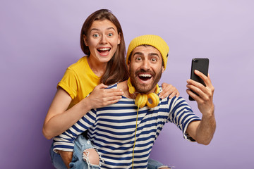Naklejka premium Pretty joyful woman rides on back of boyfriend, take selfie, have glad expressions, isolated on purple background. Cheerful nice couple on honey moon, photograph themselves on smart phone device