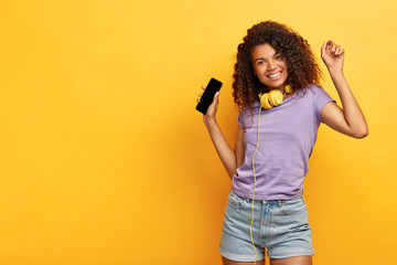 Cheerful attractive girl with curly hair, dark skin, enjoys cool songs downloaded from online website, wears headphones, holds modern smart phone, moves in music rhythm over yellow studio wall