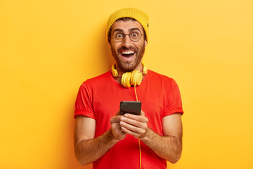 Indoor shot of optimistic Caucasian man downloads new app for listening music on mobile phone,...