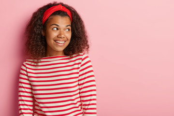Headshot of beautiful woman has no make up, curly hair, focused aside, plans how to make surprise for lover, wears red headband and striped turtleneck sweater, being in good mood after date.