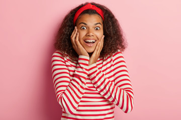 Photo of excited woman opens eyes from happiness, being pleasantly shocked, touches both cheeks, wears headband, red and white striped jumper, models indoor. People, emotions, facial expressions